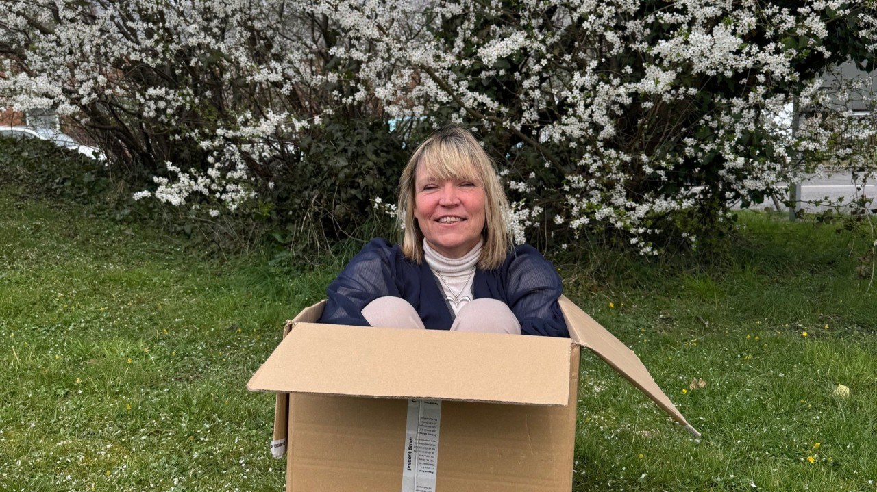 Sarah Taylor Phillips sitting in a cardboard box on a lawn under blossom, laughing.