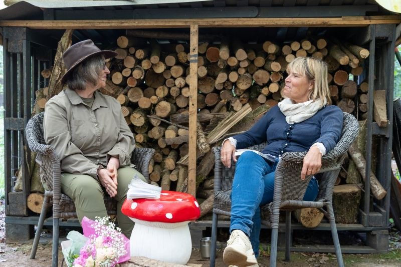 Sarah Taylor Phillips in conversation with someone outdoors, the two of them sitting in chairs in front of a log store.