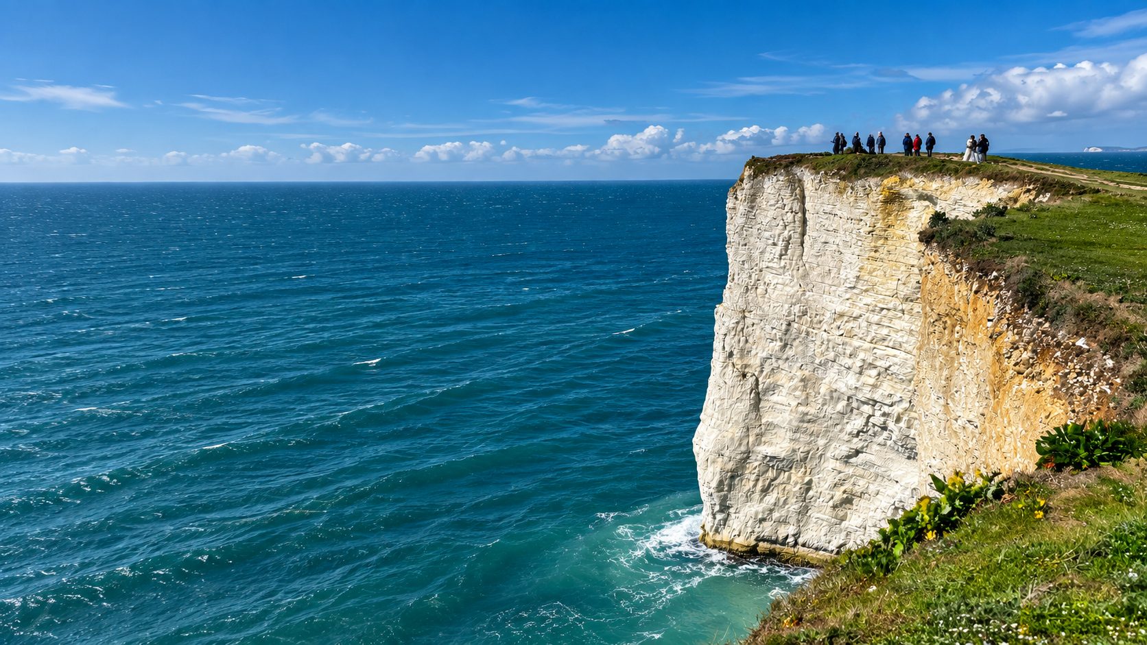 A group of walkers standing on a headland above a sheer white chalk cliff.